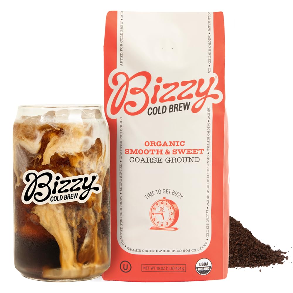 A matte black tumbler of black cold brew over ice beside a stainless cold brew pitcher, coarse ground coffee, hazelnuts, and caramel on an oak counter, illustrating a smooth low-acid coffee for cold brew.